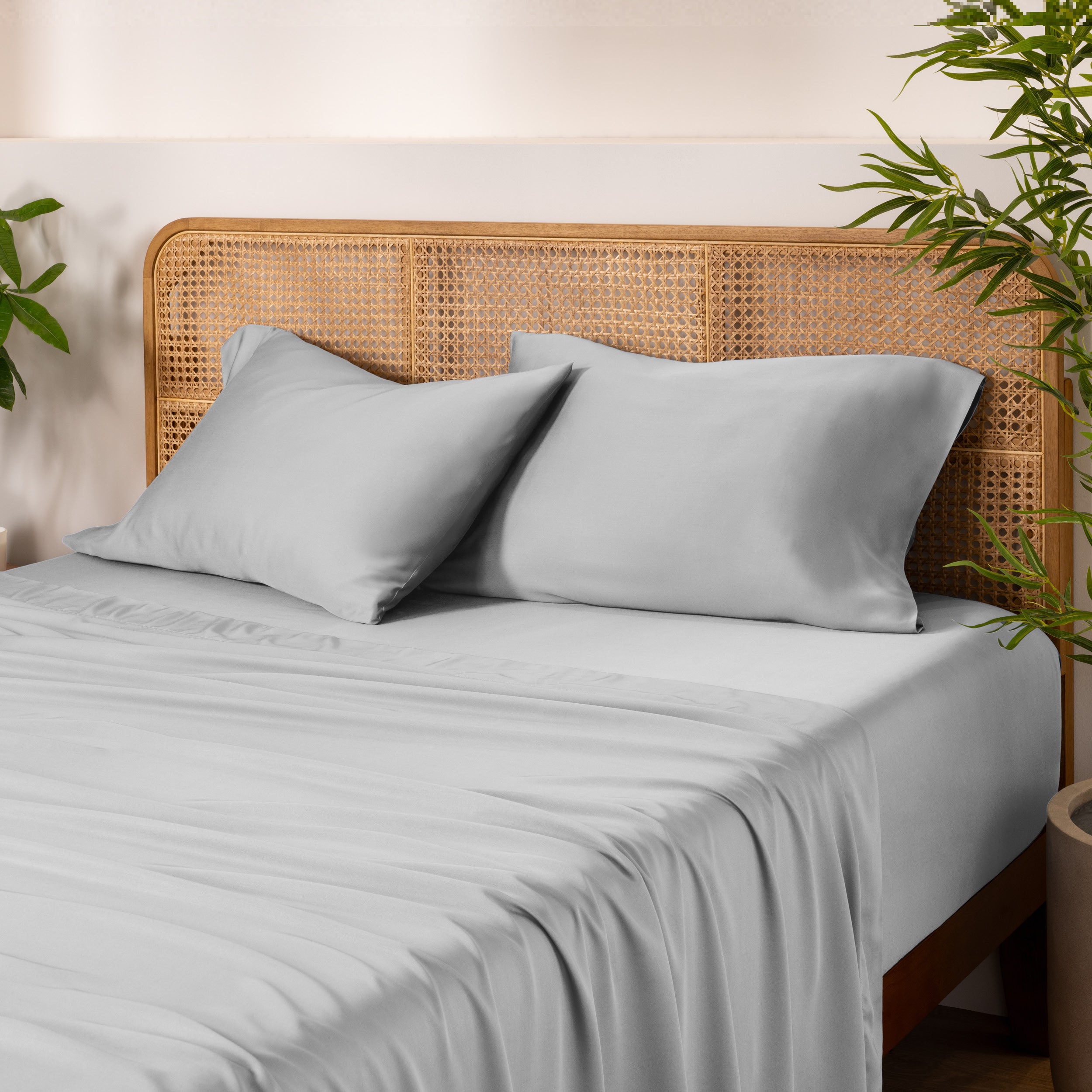Bamboo bed sheets and pillows on a bed with a wicker headboard and plants in the background.