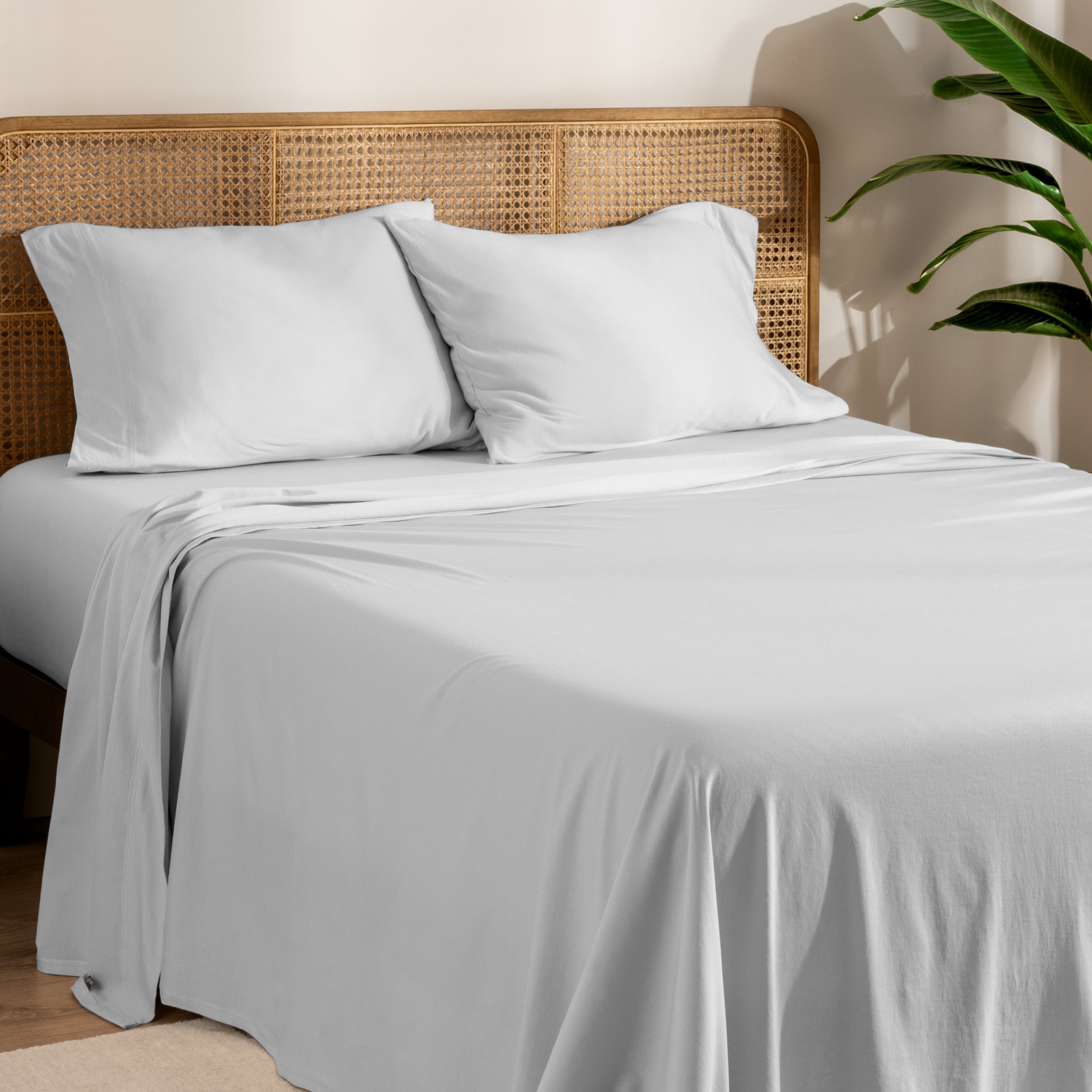 Organic cotton jersey bed sheets on a bed with a wicker headboard and plant in the background