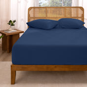 Bed with fitted sheet and pillows in a room with curtains and a plant.