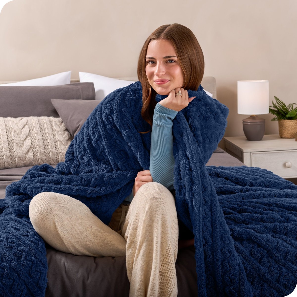 A smiling woman is sitting on the end of a bed with a sherpa blanket wrapped around her