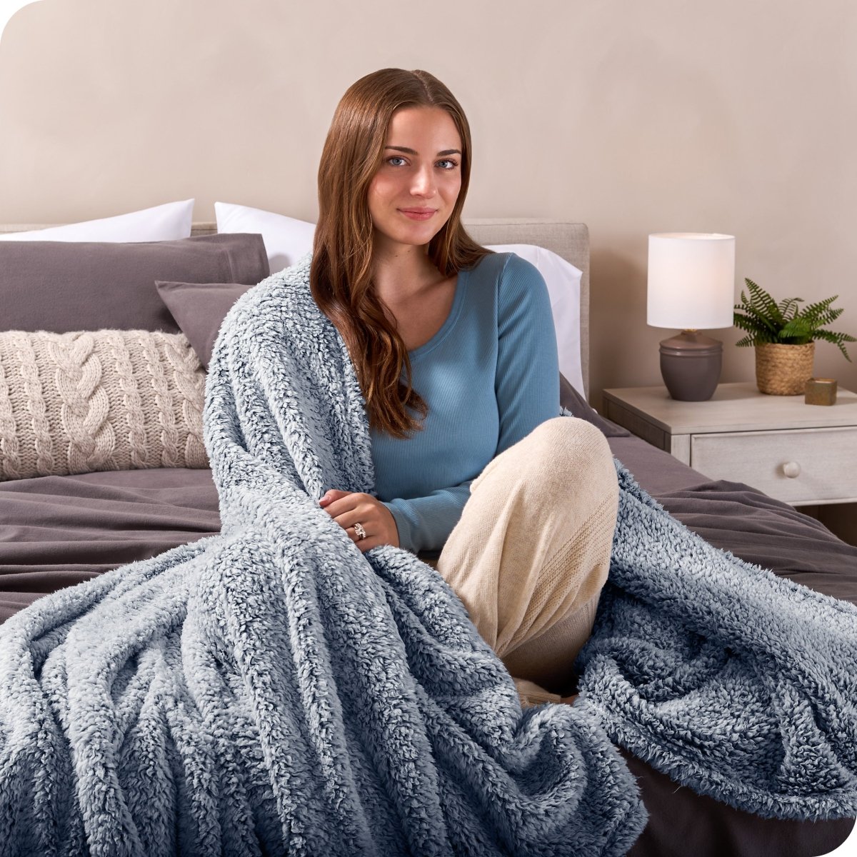 A smiling woman is sitting on the end of a bed with a sherpa blanket wrapped around her