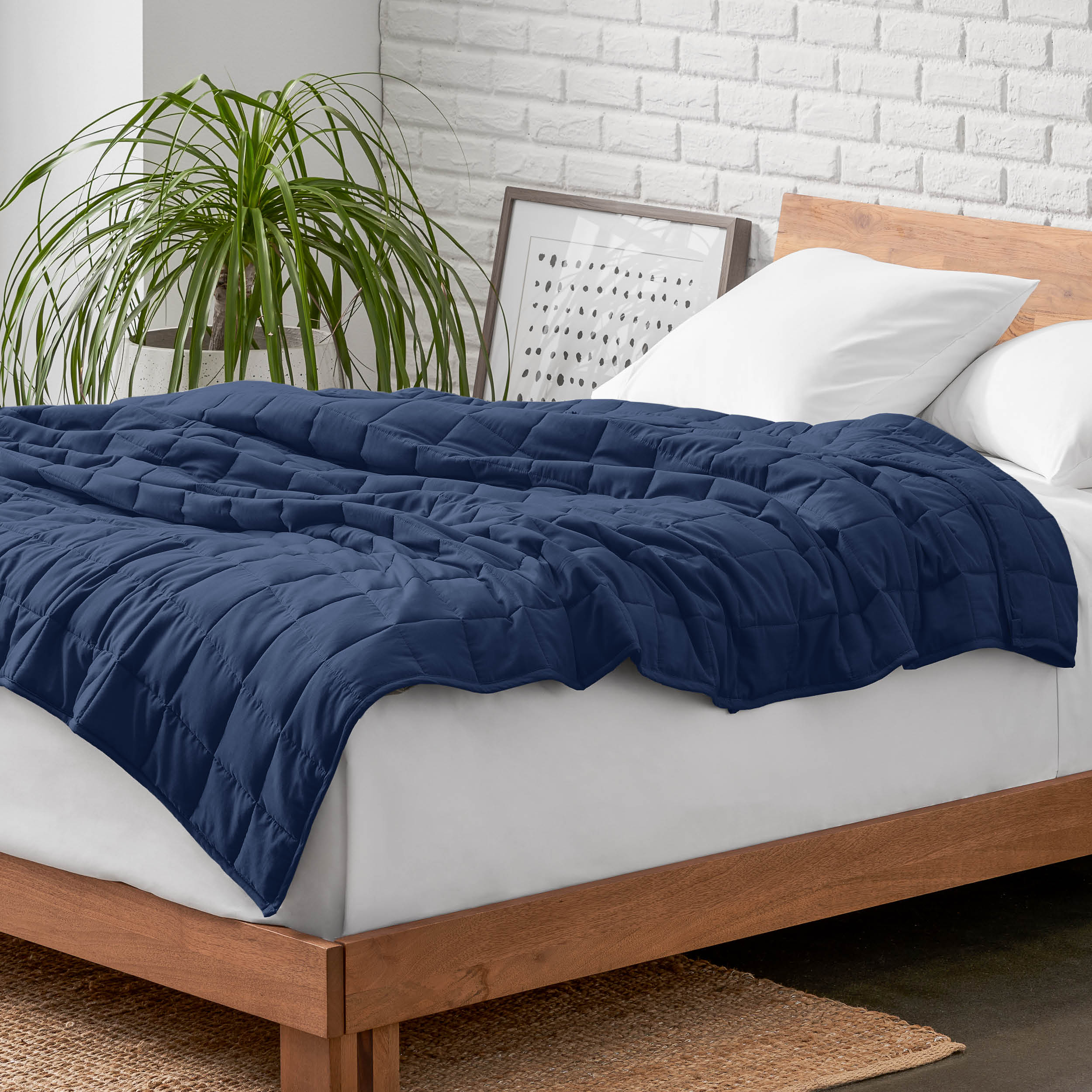 Weighted blanket on a bed with white pillows and a wooden headboard in a room with a plant and brick wall.