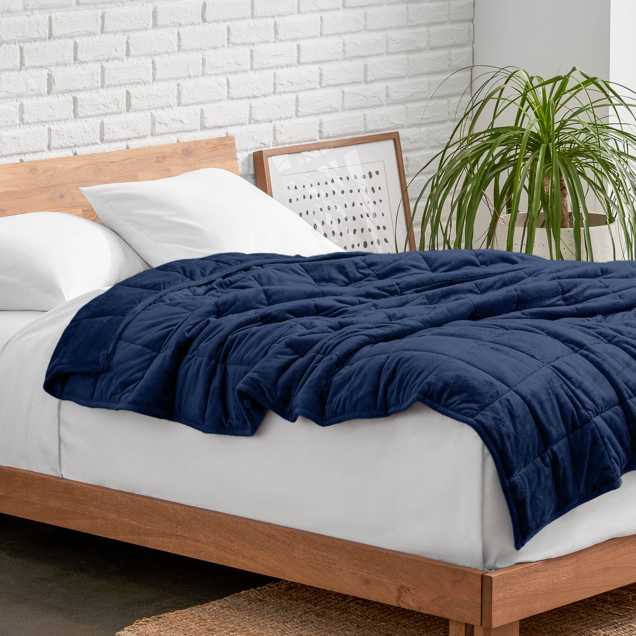 Weighted blanket on a bed with white pillows and a wooden headboard in a room with a plant and brick wall