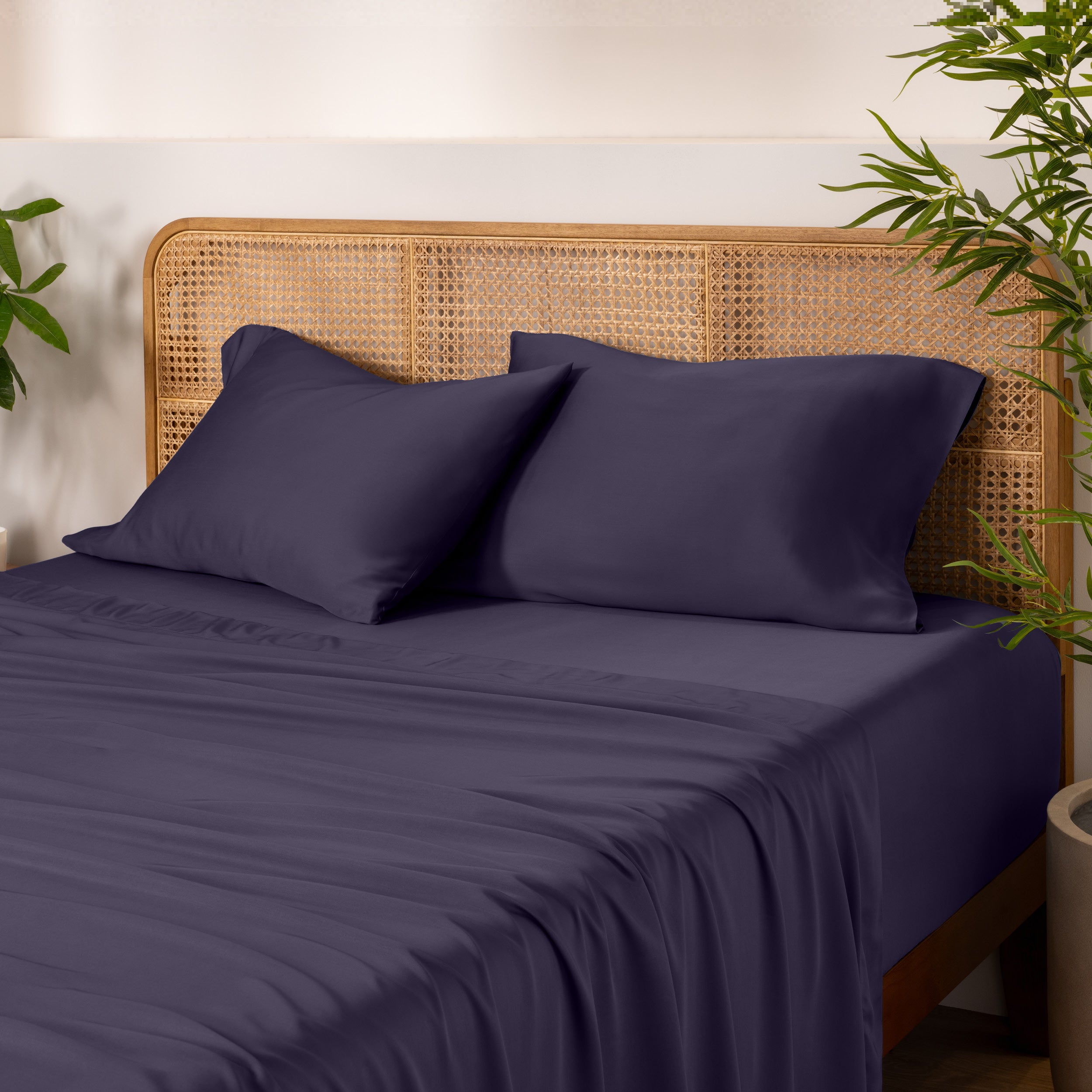 Bamboo bed sheets and pillows on a bed with a wicker headboard and plants in the background.
