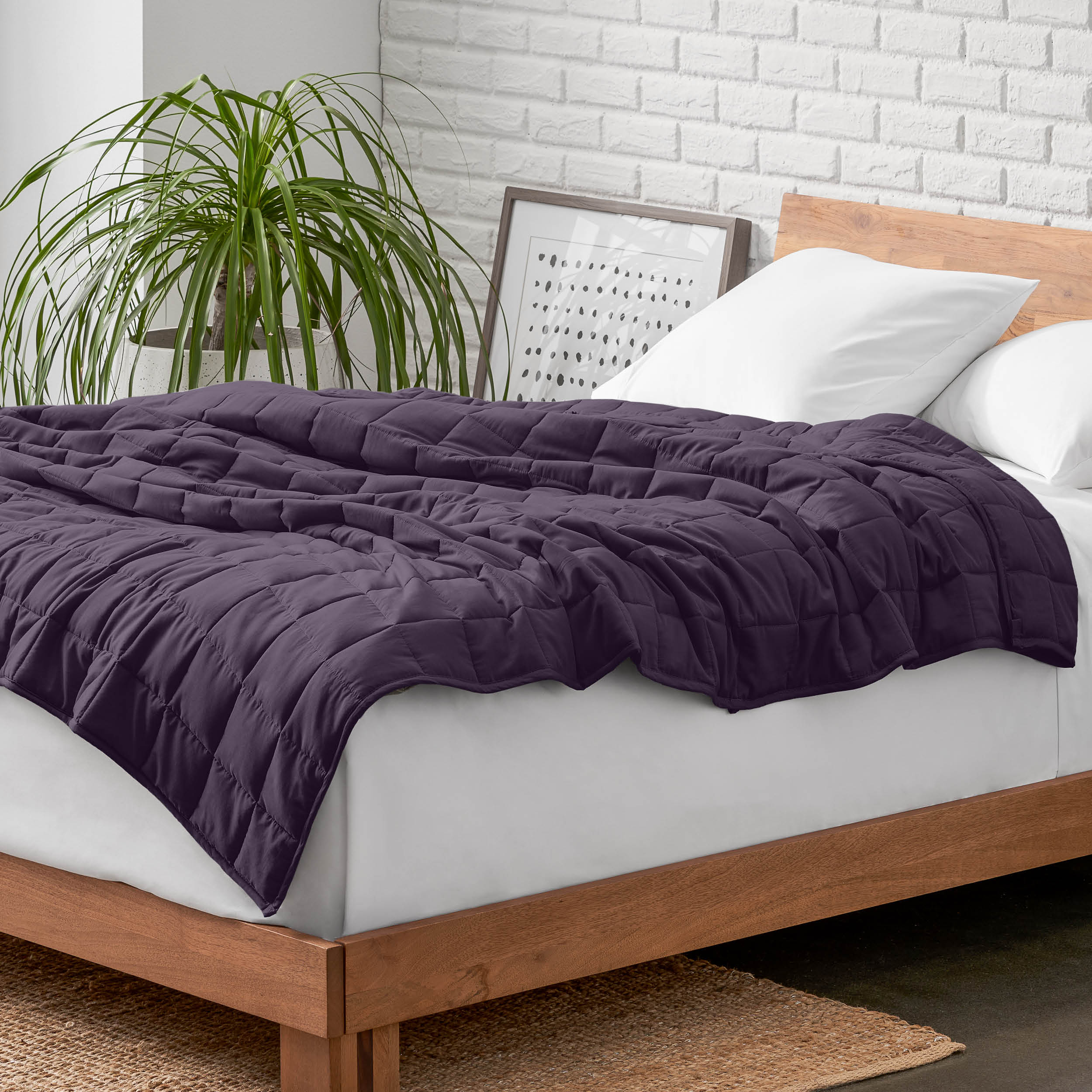Weighted blanket on a bed with white pillows and a wooden headboard in a room with a plant and brick wall.