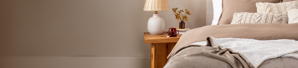 Bedroom scene with neutral tan bedding, nightstand, and lamp, collection banner