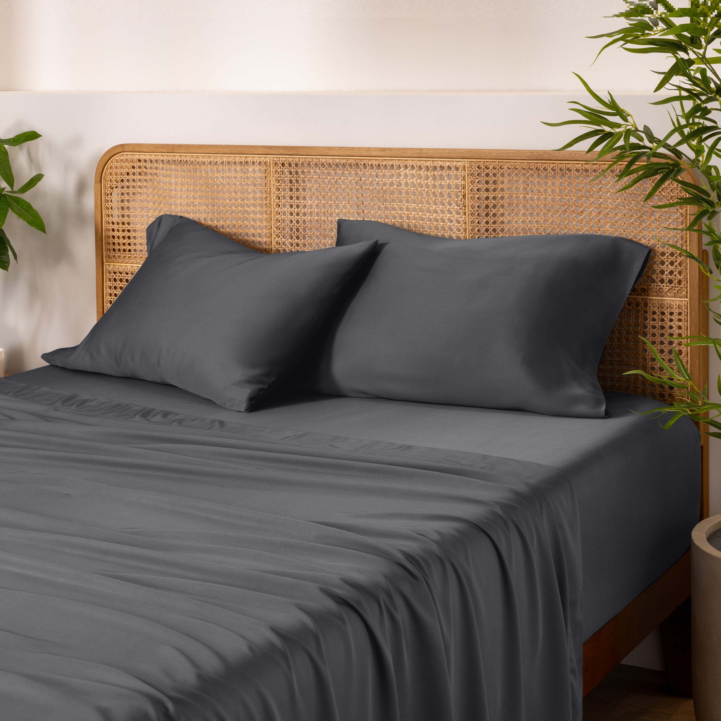 Bamboo bed sheets and pillows on a bed with a wicker headboard and plants in the background.