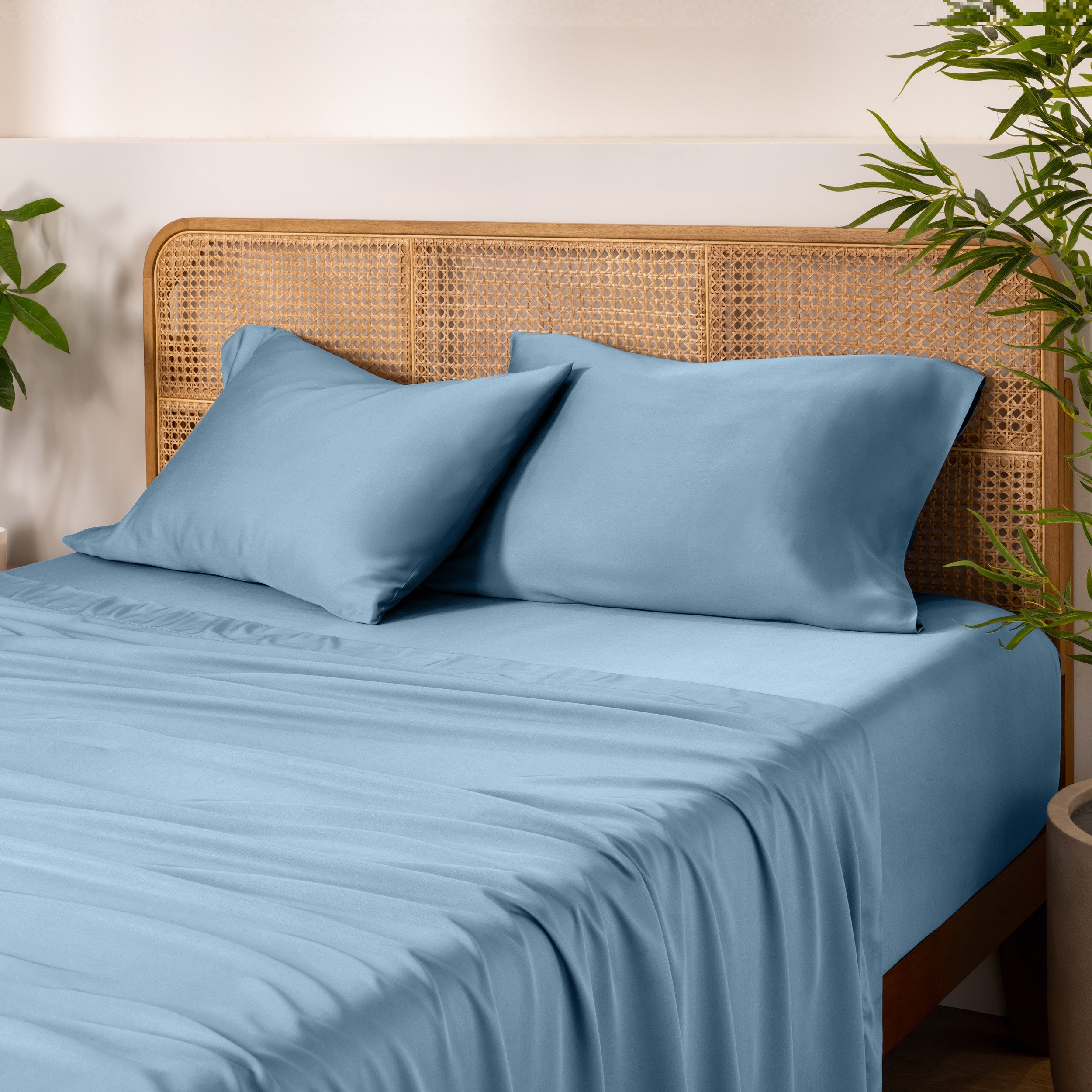 Bamboo bed sheets and pillows on a bed with a wicker headboard and plants in the background.
