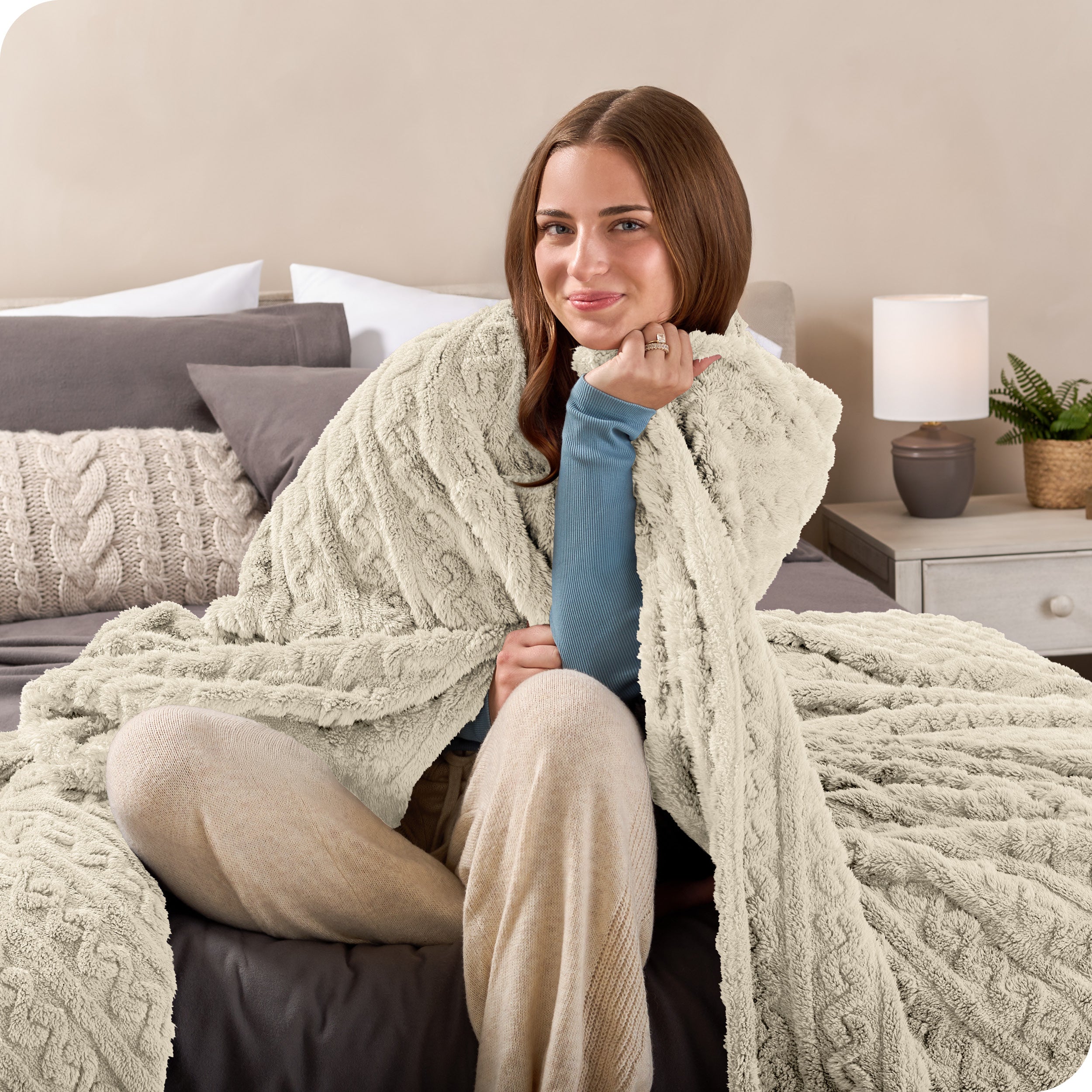 A smiling woman is sitting on the end of a bed with a sherpa blanket wrapped around her