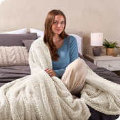 A smiling woman is sitting on the end of a bed with a sherpa blanket wrapped around her