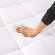 A woman is sitting on a mattress which has a white mattress pad on it.