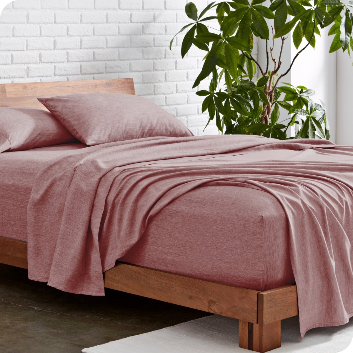 Bed with heather sheets and wooden frame against a white brick wall with a plant.