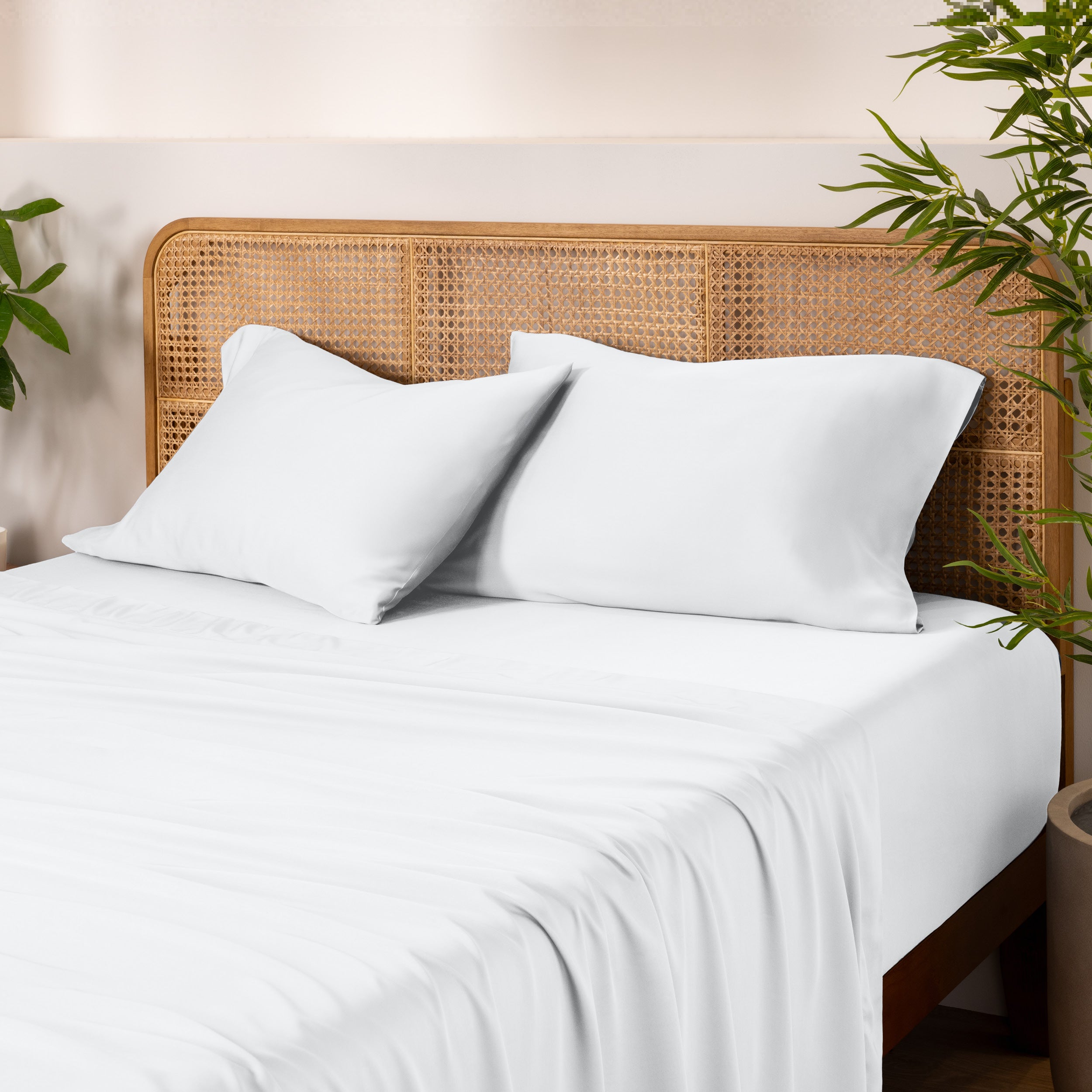 Bamboo bed sheets and pillows on a bed with a wicker headboard and plants in the background.