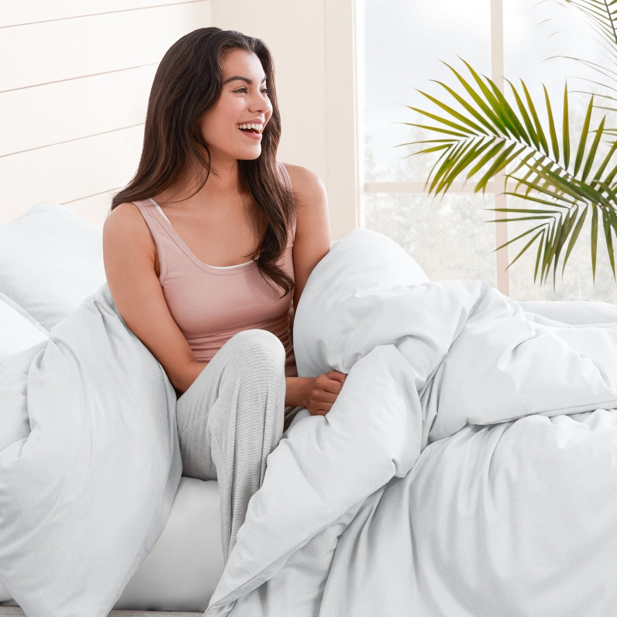 Woman sitting on a bed under duvet cover with a plant in the background