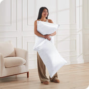 A woman is standing and holding 2 pillows covered with silk pillowcases