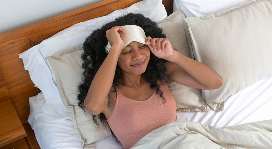 A woman reclines in bed and is in process of taking off her sleep mask.  It is daytime, and she is smiling, ready for a productive day.