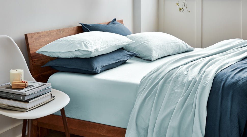 A room scene with a bed that is dressed in blue. The walls are white there is a chair next to the bed with books on it.