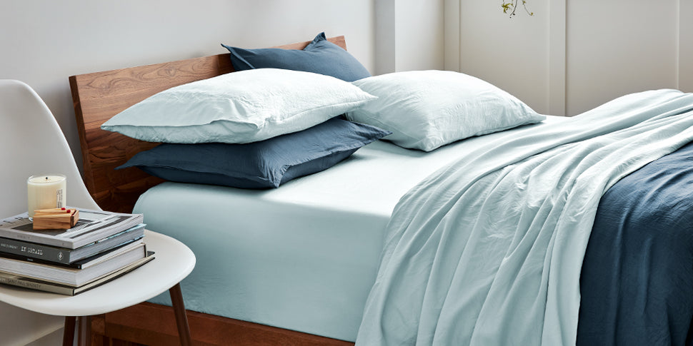 A room scene with a bed that is dressed in blue. The walls are white there is a chair next to the bed with books on it.