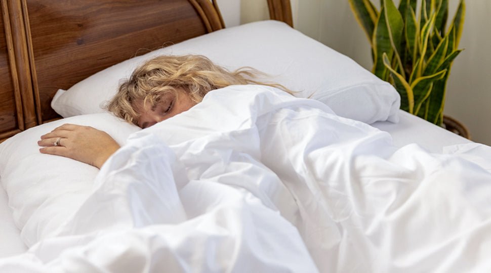 A young woman is lying on a bed. Her head is resting on a pillow and she has a white duvet covering her.