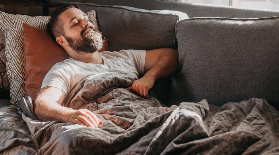 A man is sleeping on a couch with two pillows behind his head and a blanket covering him.