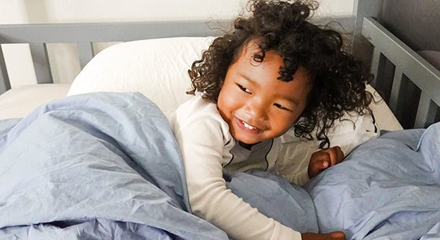 A little girl with curly hair snuggles up in a crib. She is smiling at someone off camera.