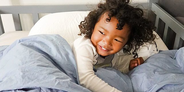 A little girl with curly hair snuggles up in a crib. She is smiling at someone off camera.