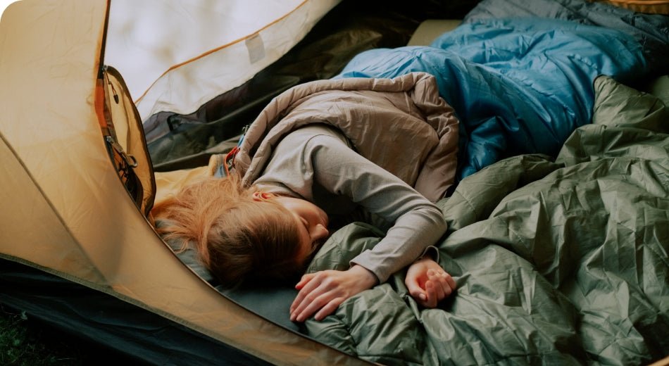 A woman sleeps inside of a sleeping bag.  She is lying on top of a sleeping pad in a tent.  Colors are natural – green, blue and tan.