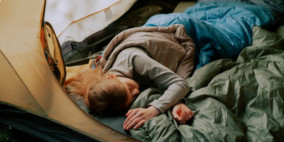 A woman sleeps inside of a sleeping bag.  She is lying on top of a sleeping pad in a tent.  Colors are natural – green, blue and tan.