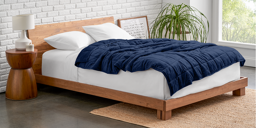 Bed with a navy minky fleece weighted blanket on a wood frame next to a plant