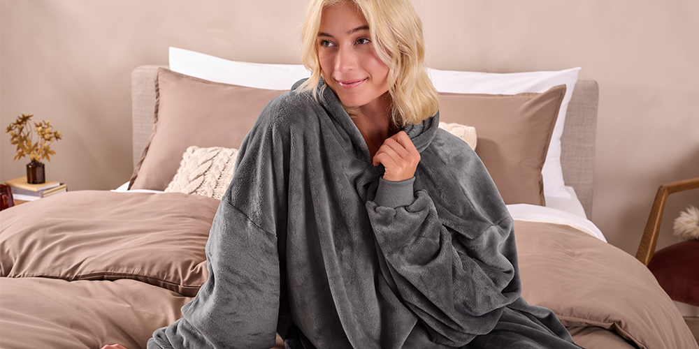 Woman on a bed wearing a gray Bare Homey Wearable Blanket (Microplush Adult) with hood up