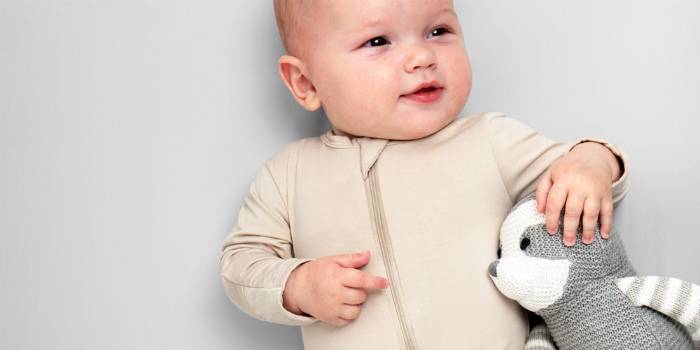 Baby sitting on a cream crib sheet with a stuffed animal