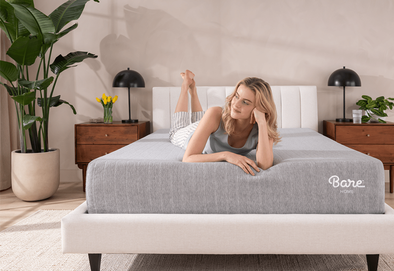 Woman lying on a bed with grey and white bedding, blog image about CertiPUR-US certified mattresses