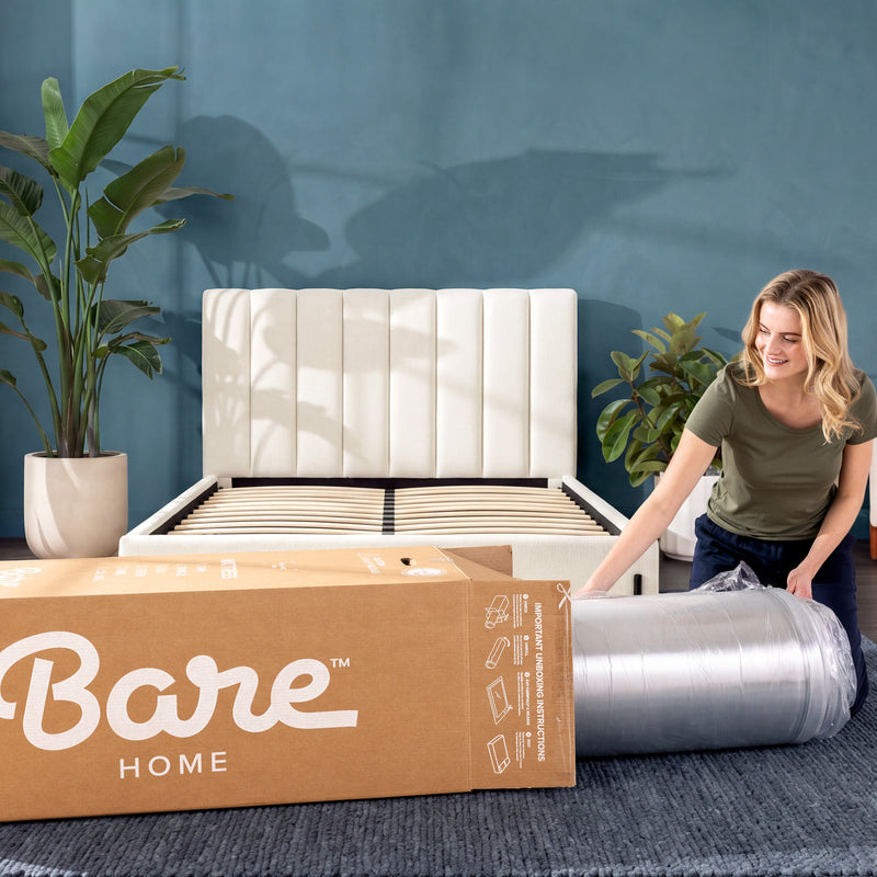 A woman unrolls a silver metal bed frame over a slatted base in a styled room with a blue wall, potted plants, and a large cardboard box labeled Bare Home in the foreground.