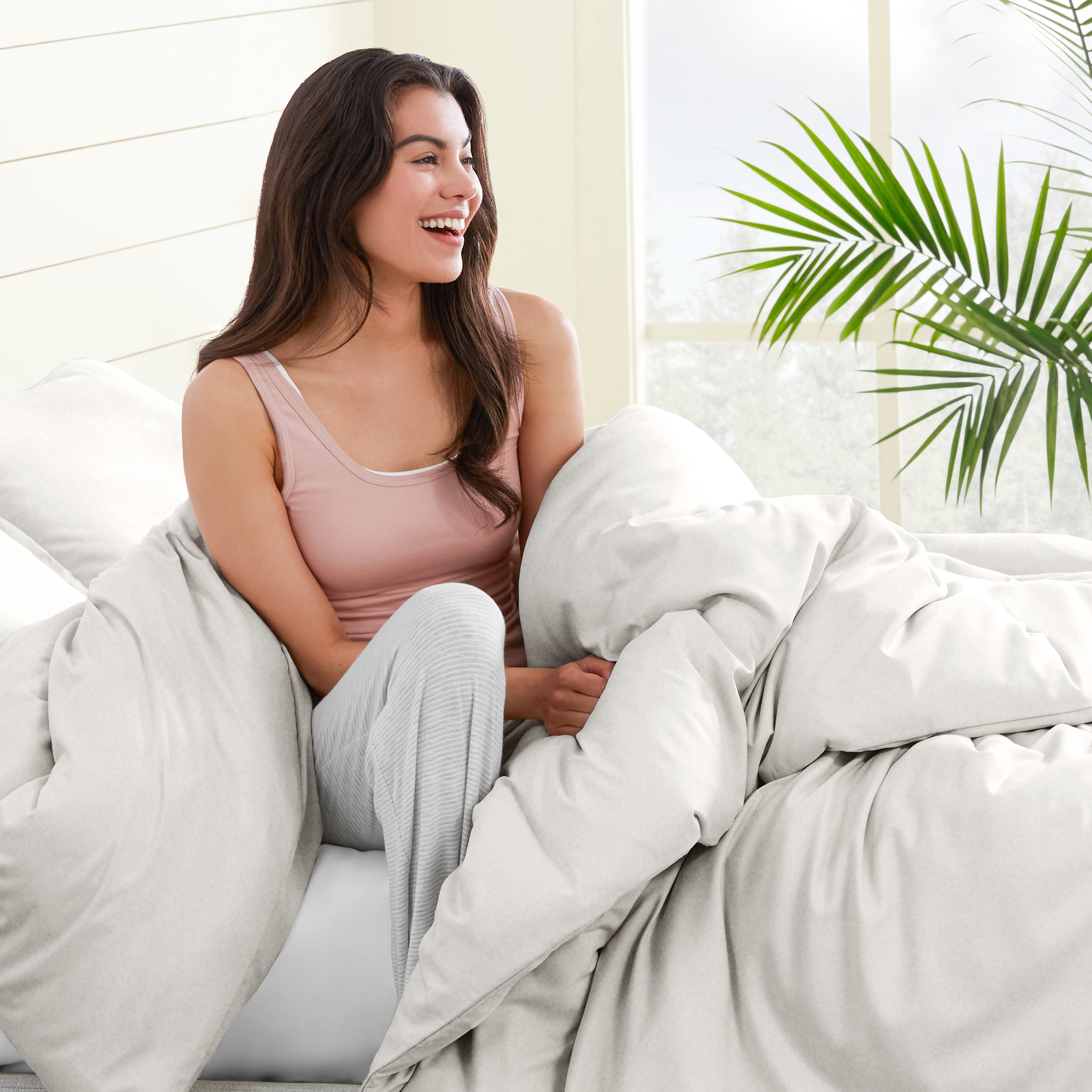 Woman sitting on a bed under duvet cover with a plant in the background