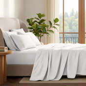 A neatly made bed with in a softly lit bedroom. A large green plant stands beside the bed, and a window with curtains is visible in the background.
