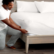Man adjusting fitted sheet on a bed with white pillows.