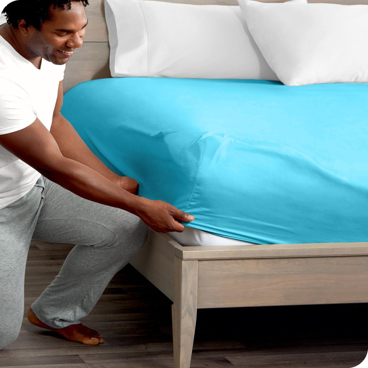 Man in white shirt and gray pants adjusting a fitted sheet on a bed.