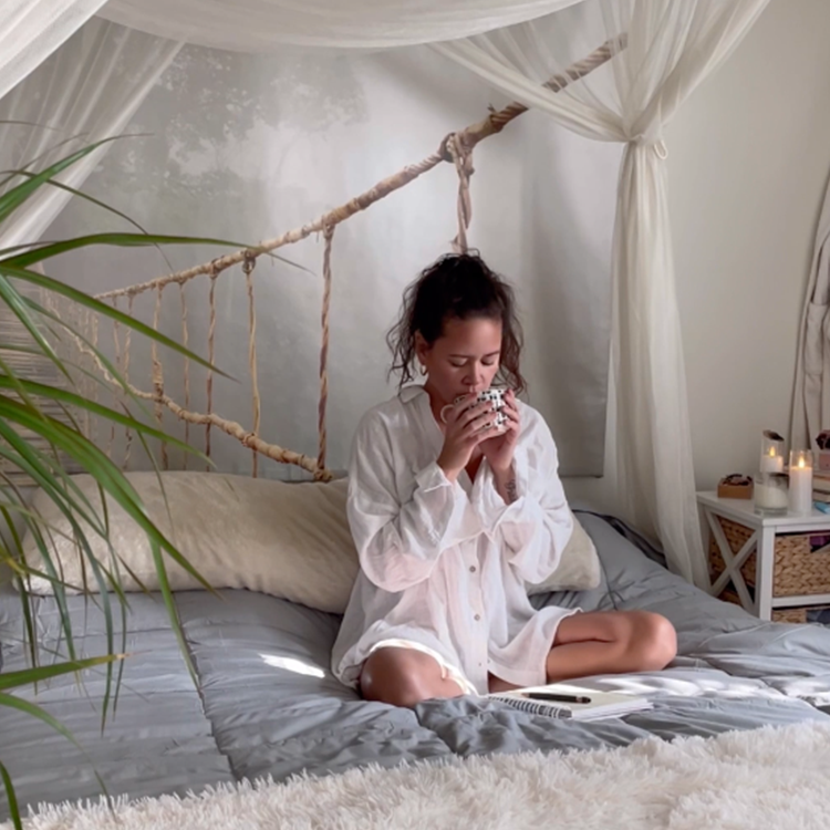 Woman sitting on a bed in a cozy room with plants and candles.