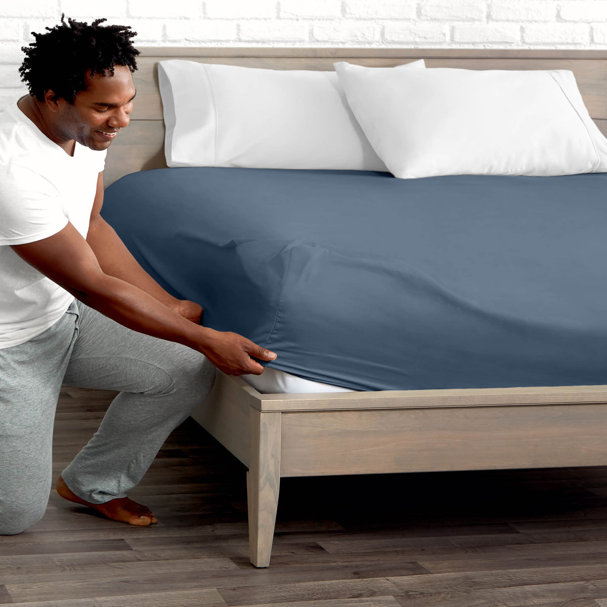 Man adjusting fitted sheet on a bed with white pillows.