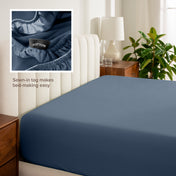 Fitted sheet on a bed with a close-up of a sewn-in tag, nightstand, lamp, and plant in a bedroom setting.