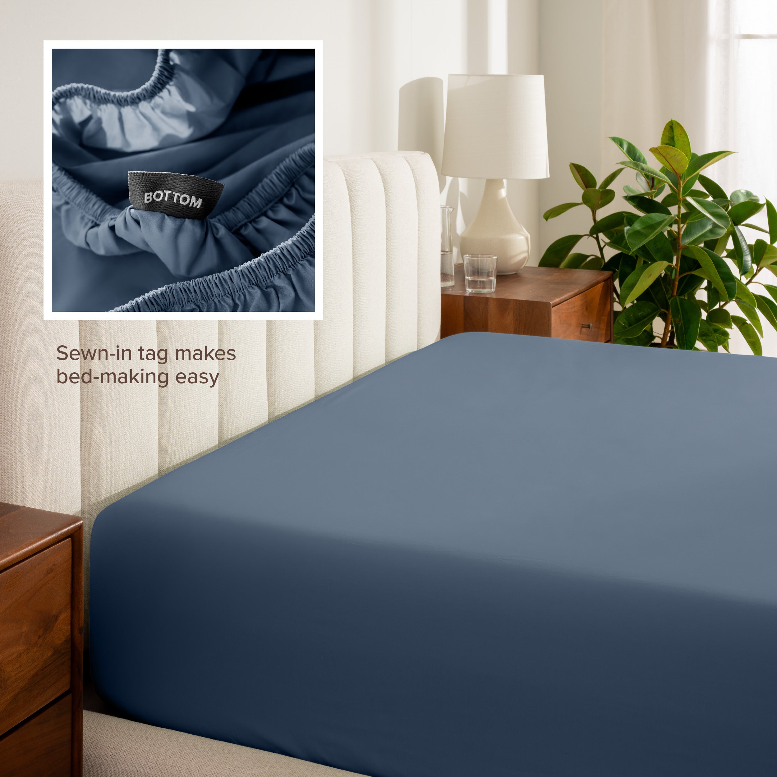 Fitted sheet on a bed with a close-up of a sewn-in tag, nightstand, lamp, and plant in a bedroom setting.