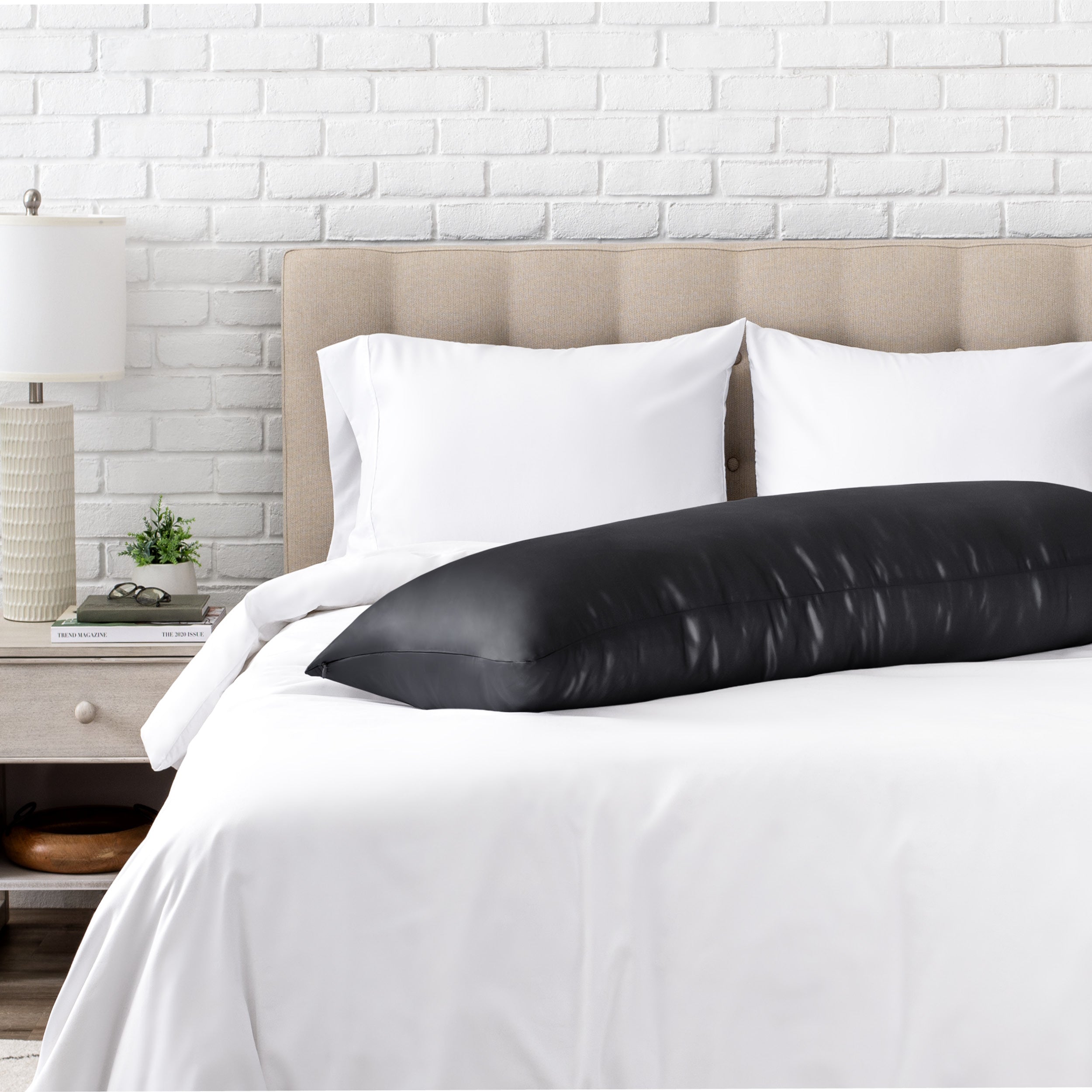 Bed with white bedding and a body pillow against a white brick wall.