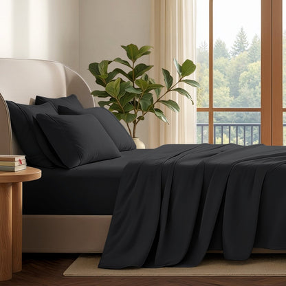 A neatly made bed with in a softly lit bedroom. A large green plant stands beside the bed, and a window with curtains is visible in the background.