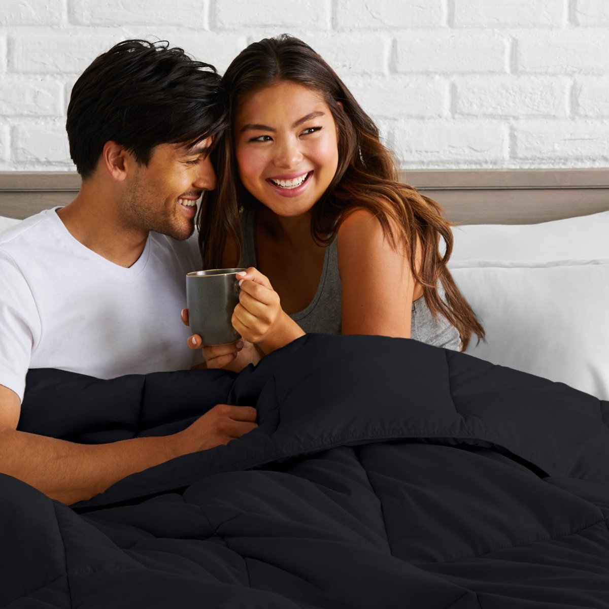 Man and woman sitting together in bed holding a mug, smiling.
