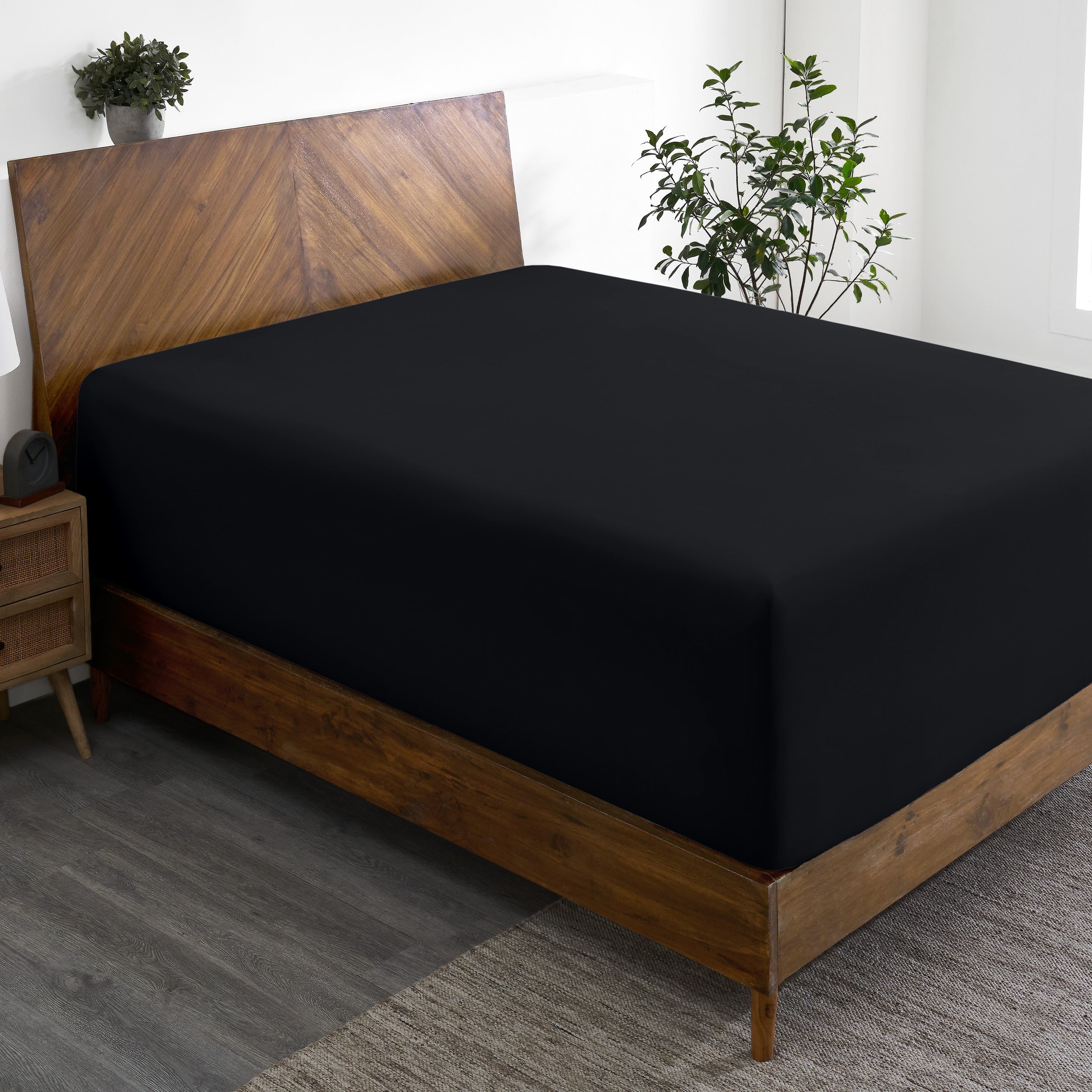 Wooden bed frame with a fitted sheet on the mattress in a room with plants and a nightstand.