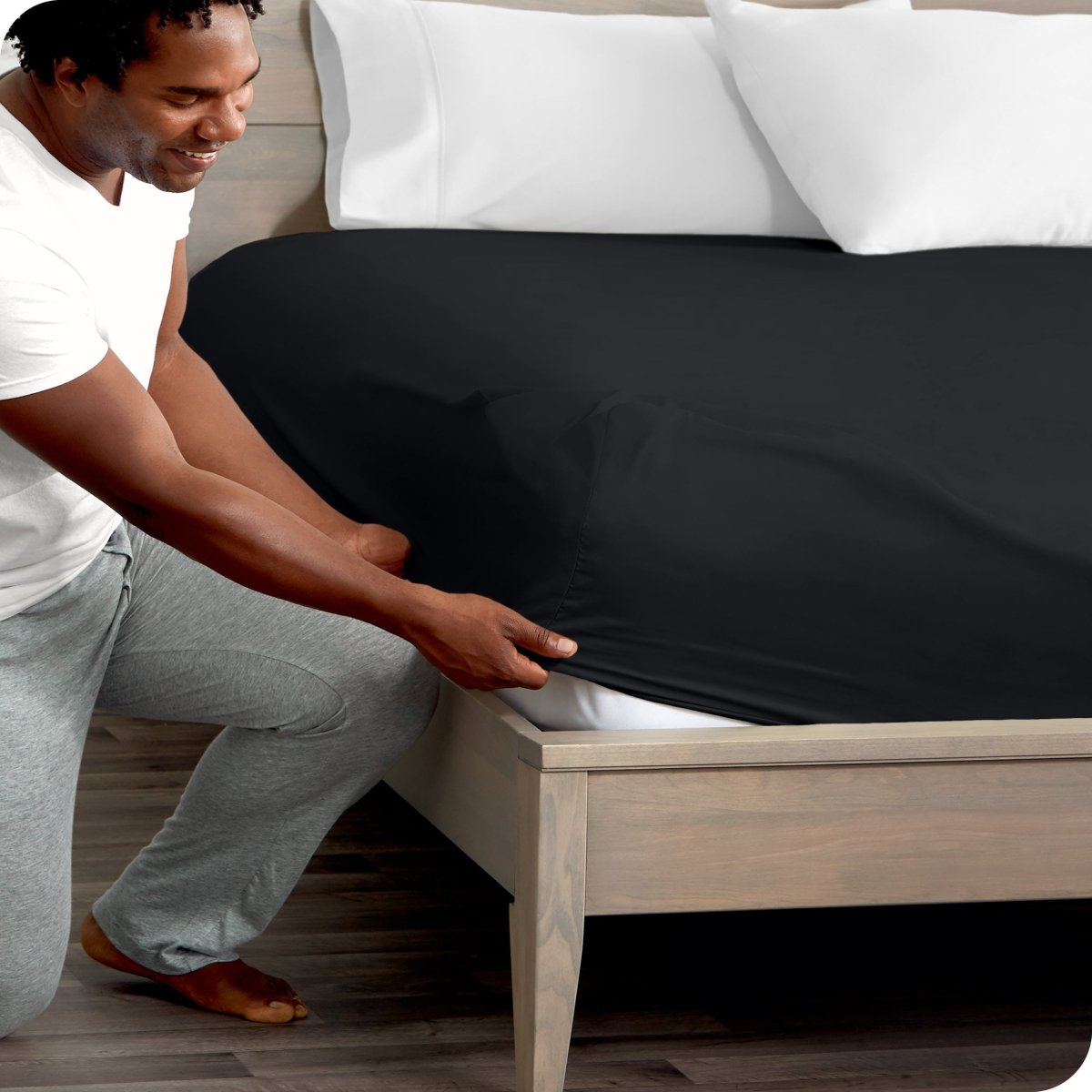 Man in white shirt and gray pants adjusting a fitted sheet on a bed.