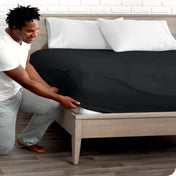 A man is kneeing and putting the corner of the fitted sheet on the mattress. The mattress is on a wooden bed frame.