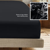 A fitted bed sheet on a bed frame with a small inset close-up of the elastic and a sewn-in tag labeled BOTTOM. White text on the sheet reads, 'Sewn-in tag makes bed-making easy.