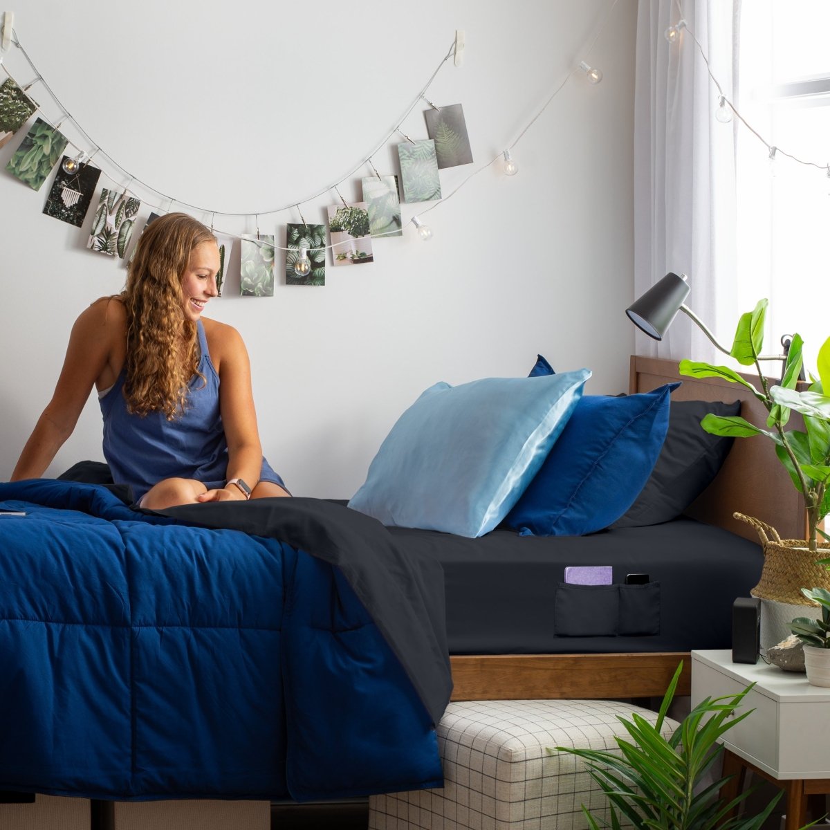 Woman sitting on a bed with blue and black bedding, surrounded by plants and decor.