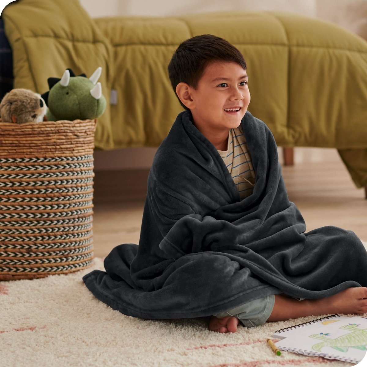 A boy is sitting on the floor of his bedroom with a microplush blanket wrapped around him